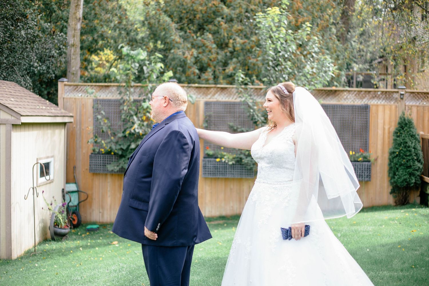 Bride in white lace wedding gown approaching father from behind for an emotional first look in backyard.