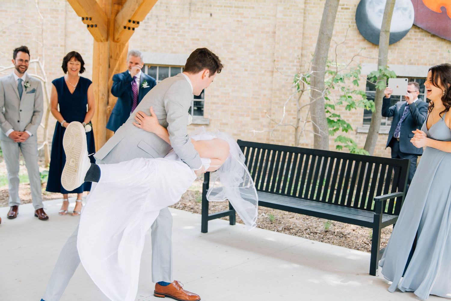 Groom dips bride dramatically during outdoor wedding first look while guests watch and smile.