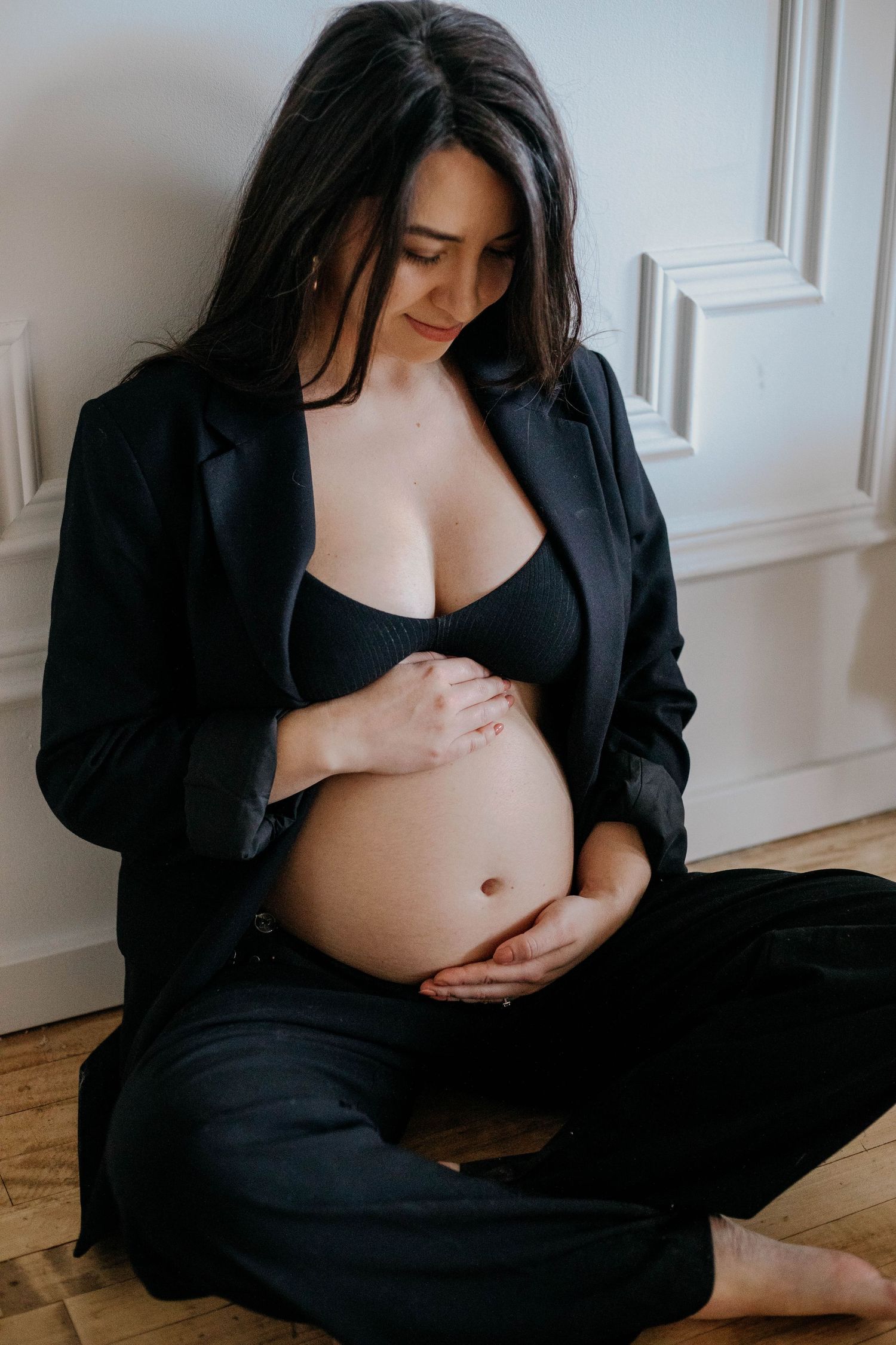 Pregnant woman in black blazer and bra sitting on floor, gently holding her baby bump while looking down.