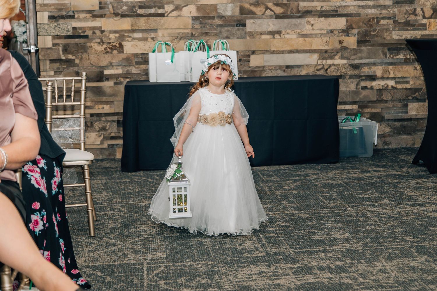 Young flower girl in white tulle dress carries lantern down wedding aisle in decorated ceremony venue.