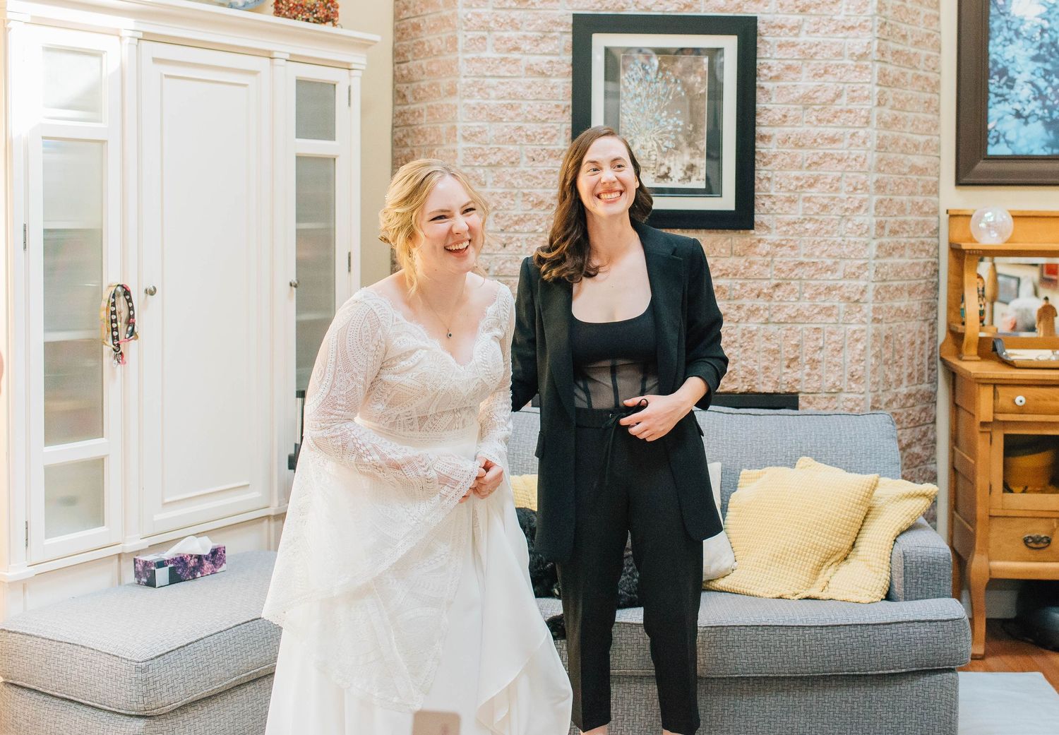 A bride in a lace dress and a woman in a black outfit smiling in a bright living room.