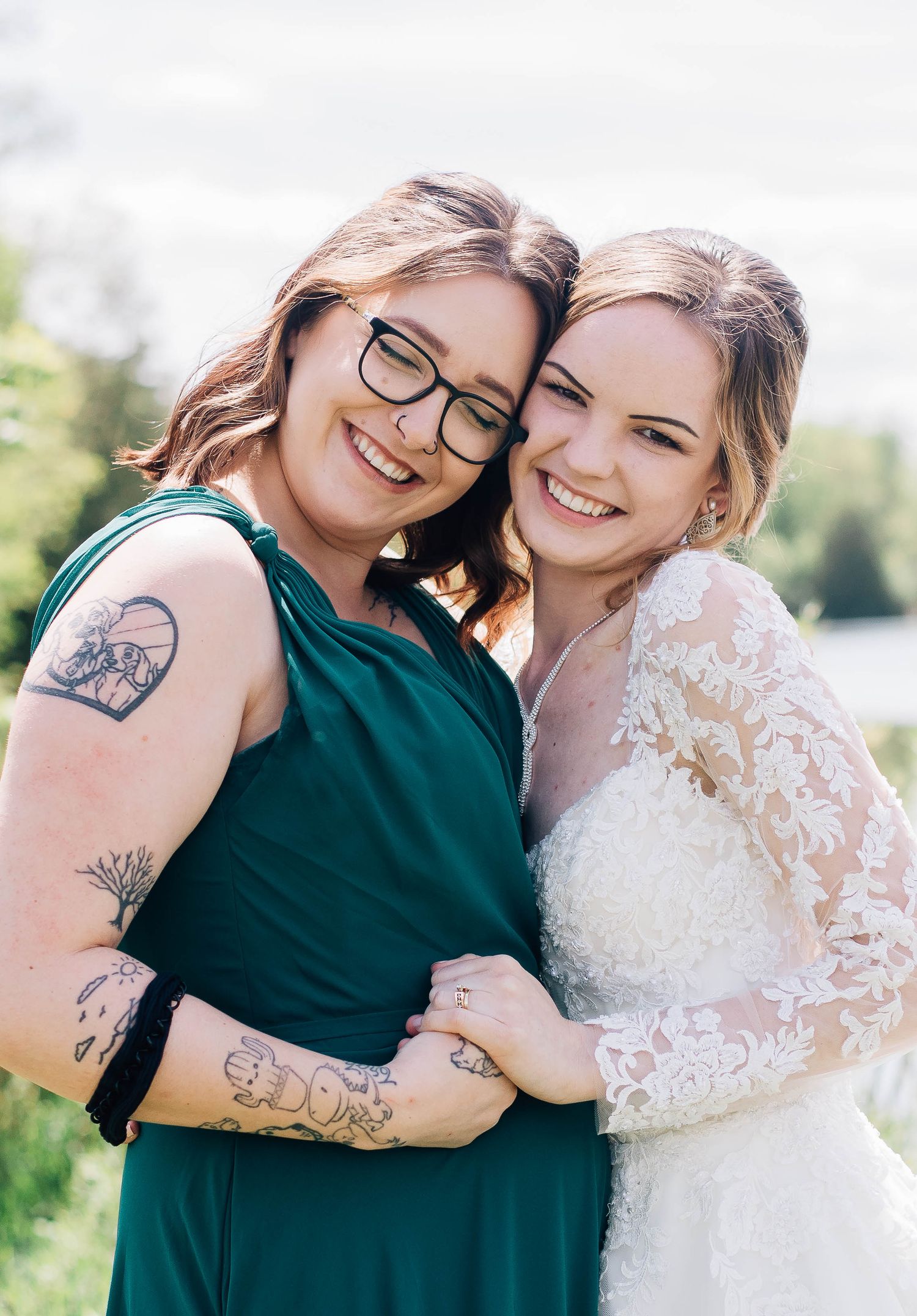 Bride in lace wedding dress smiling with bridesmaid in emerald green dress outdoors.