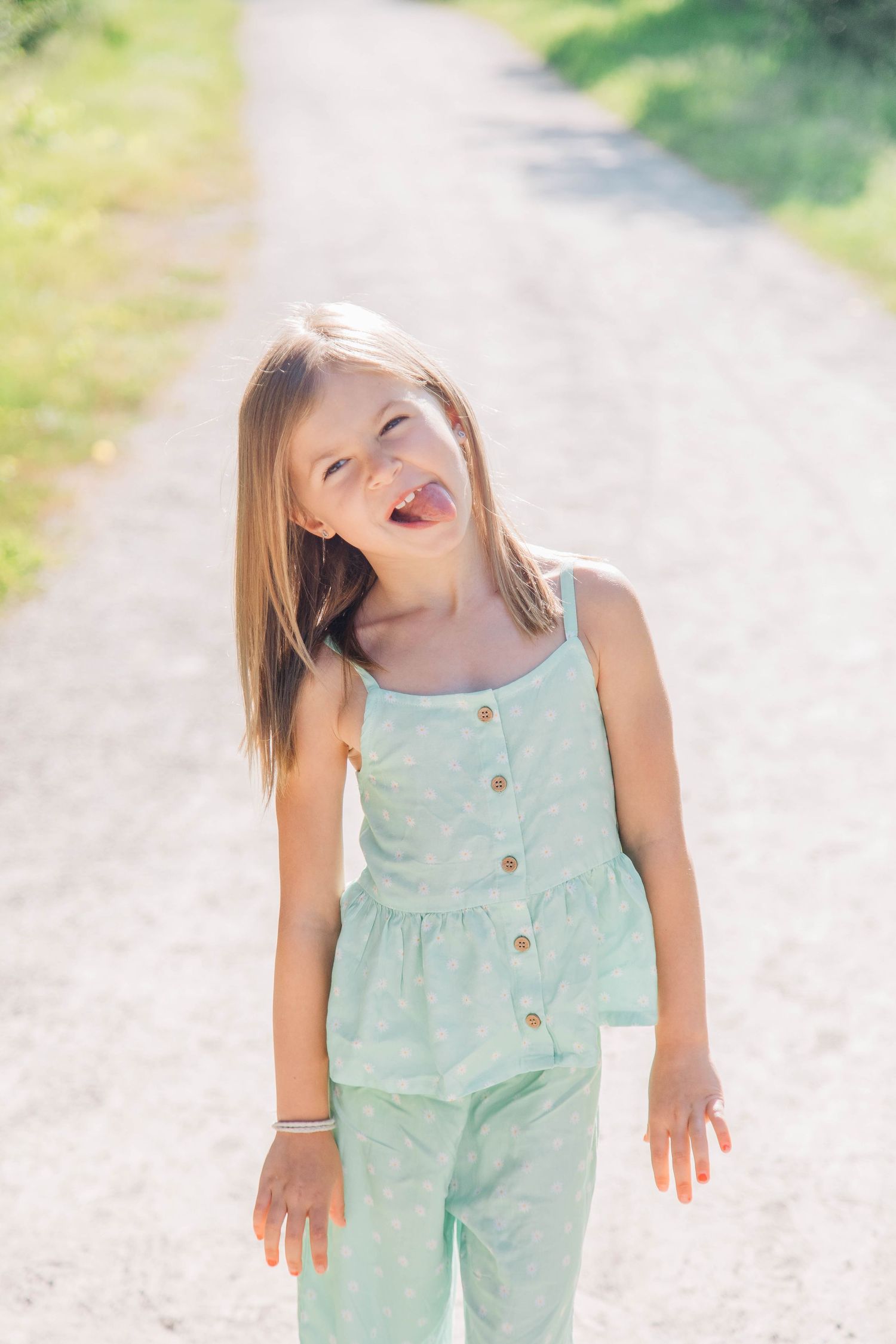 Young girl in a mint green jumpsuit sticking her tongue out playfully on a sunny outdoor path.