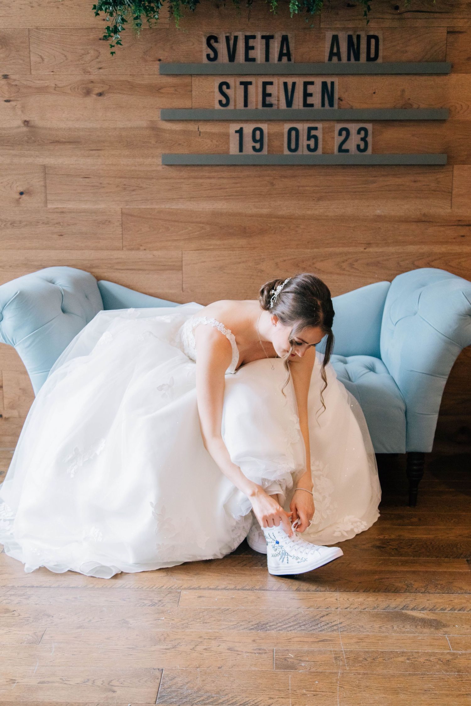 Bride in white ballgown tying her sneakers on a blue velvet sofa beneath a Sveta and Steven sign.