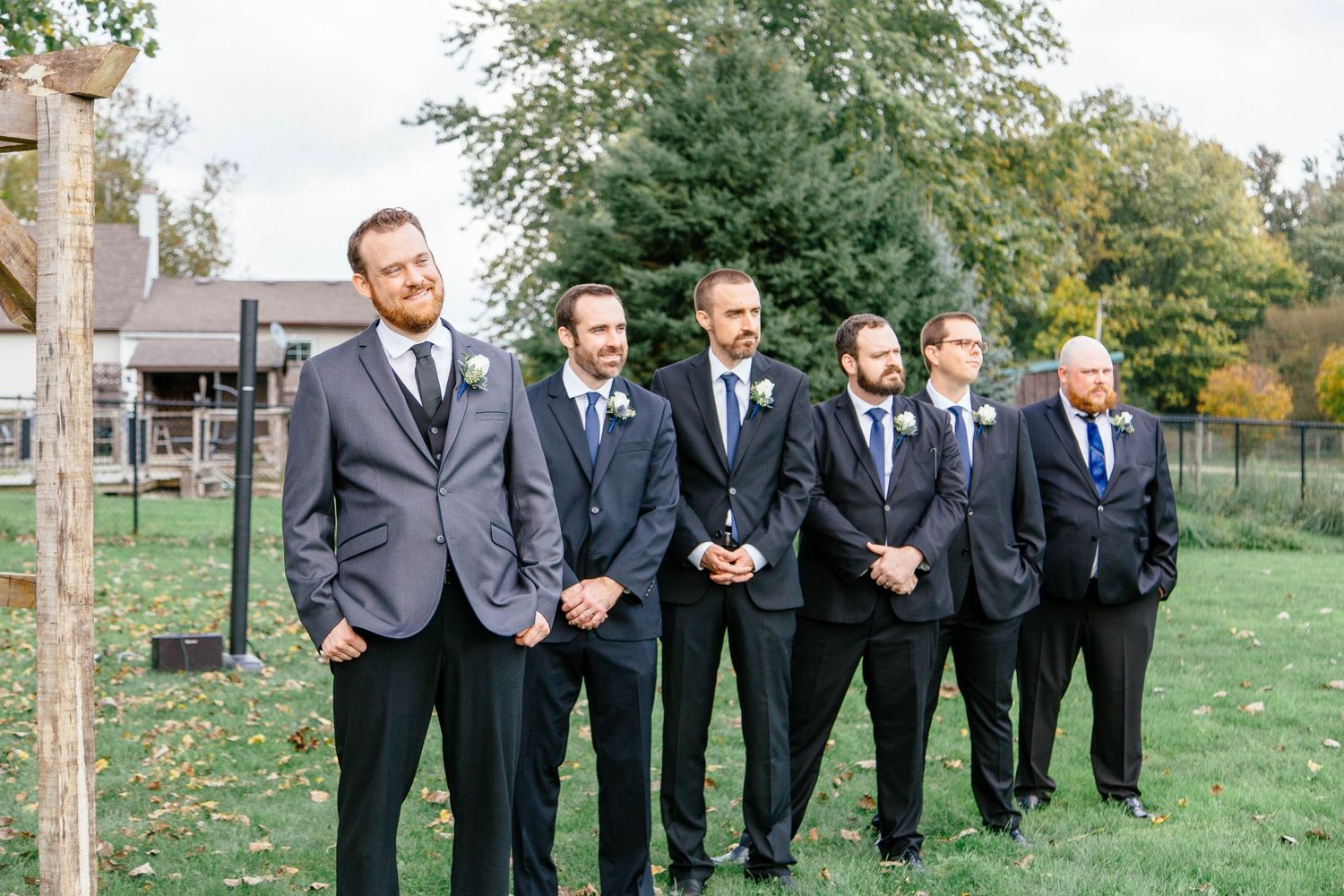 Groom in gray suit stands with groomsmen in navy suits outdoors at a fall wedding ceremony.