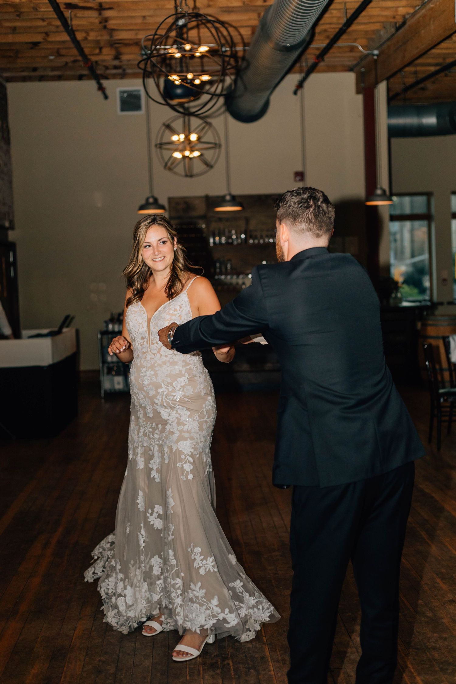 Bride in lace gown twirls with groom during first dance under ornate chandelier at wedding reception.