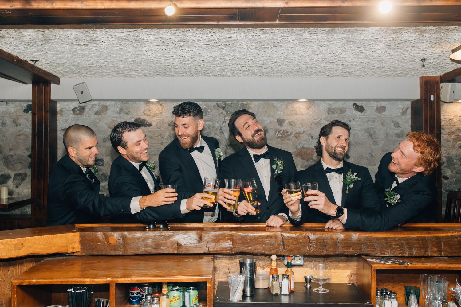 Groom and groomsmen in tuxedos toasting drinks together at a rustic bar inside a hotel in Paris Ontario.