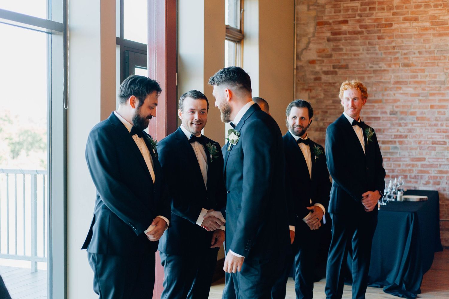 Groom in black tuxedo greets smiling groomsmen before wedding ceremony in elegant venue.