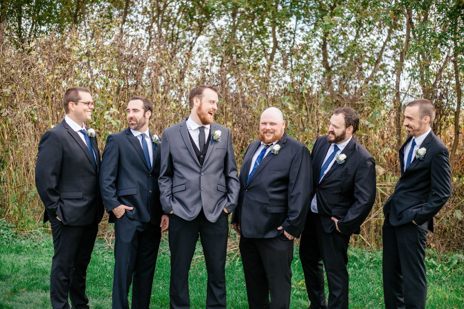 Groom with groomsmen in navy suits and blue ties posing outdoors near autumn foliage at wedding.