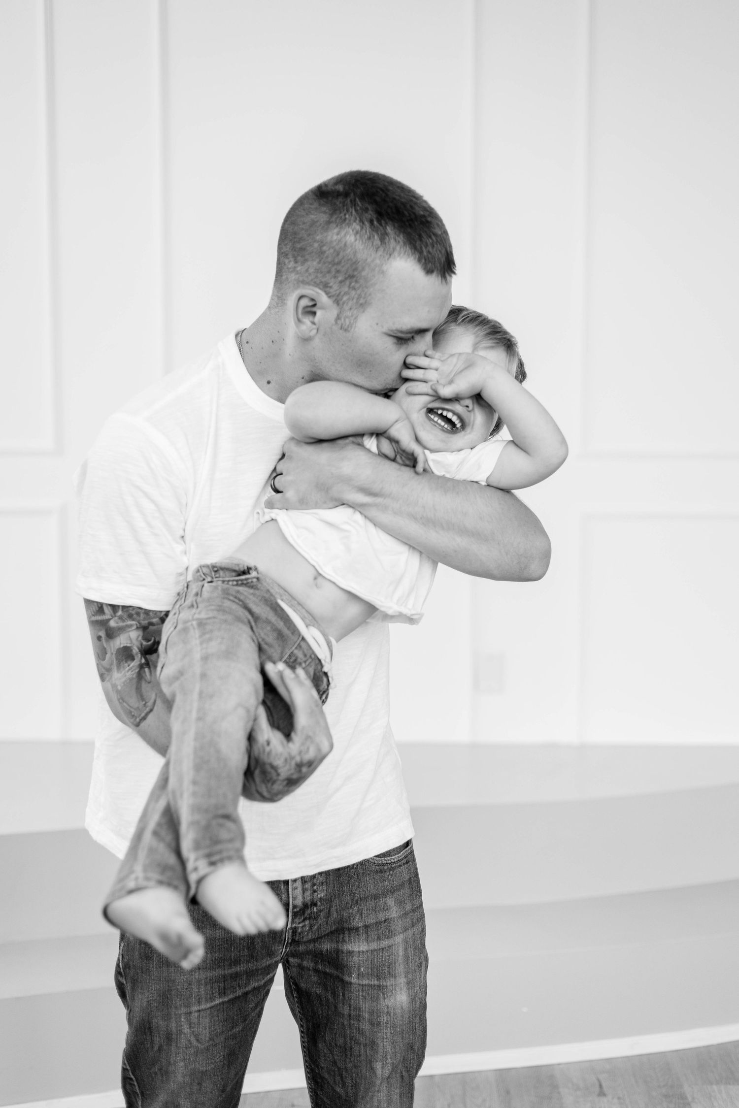 Father kissing laughing toddler in black and white family photo.