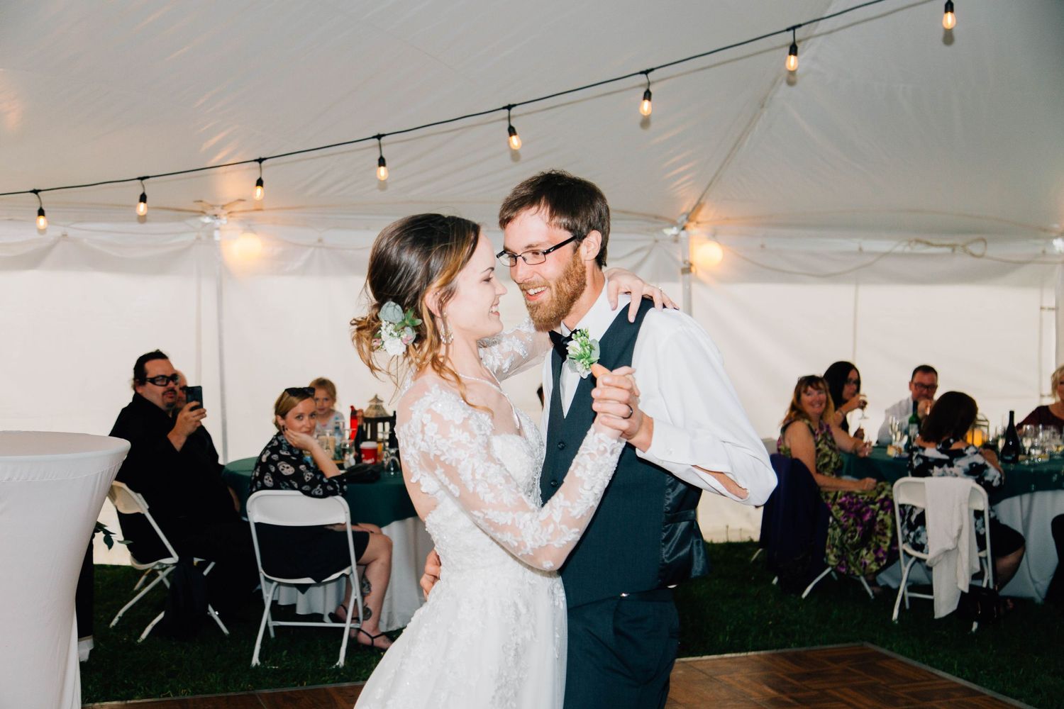 Newlyweds share romantic first dance under string lights in elegant wedding reception tent.