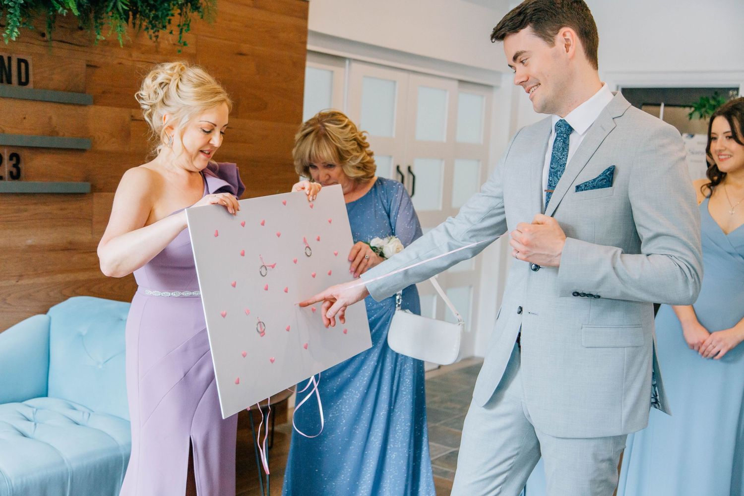 Groom in gray suit examines decorative wedding poster held by bridesmaid in lavender dress at indoor venue.