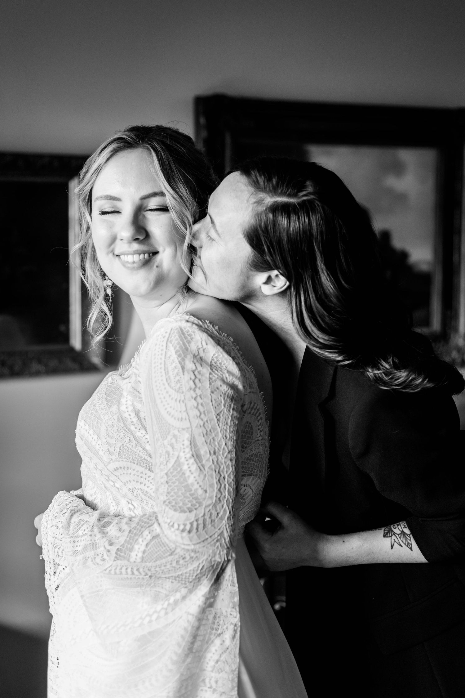A black and white photo of a bride smiling as her partner affectionately kisses her cheek before their wedding.