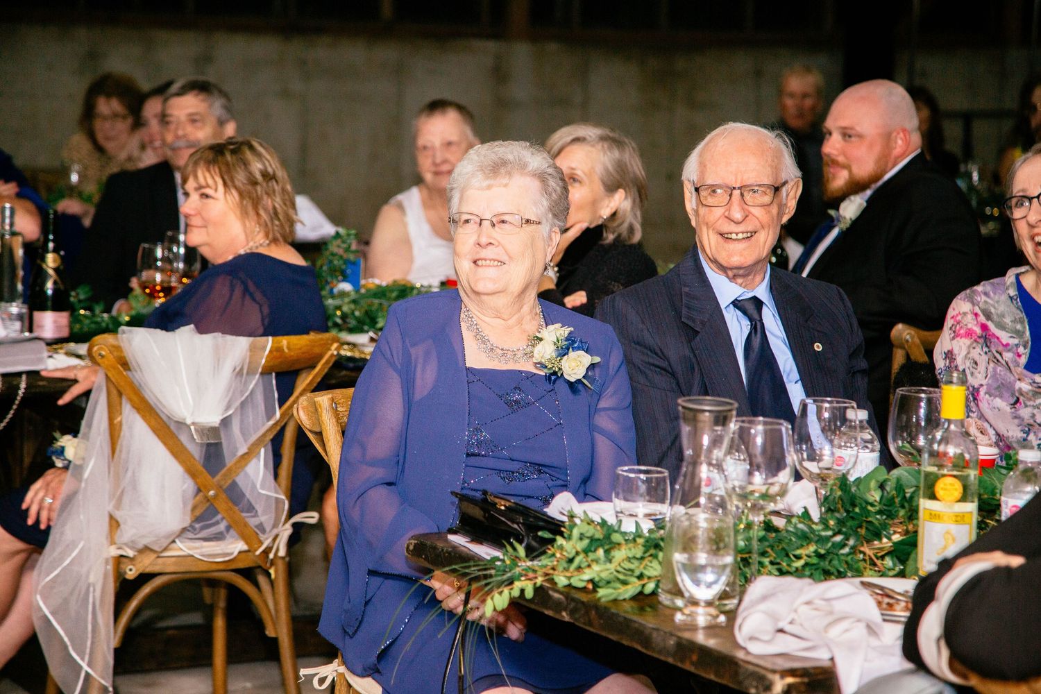 Elderly couple in navy blue attire smiling warmly at wedding reception dinner table.