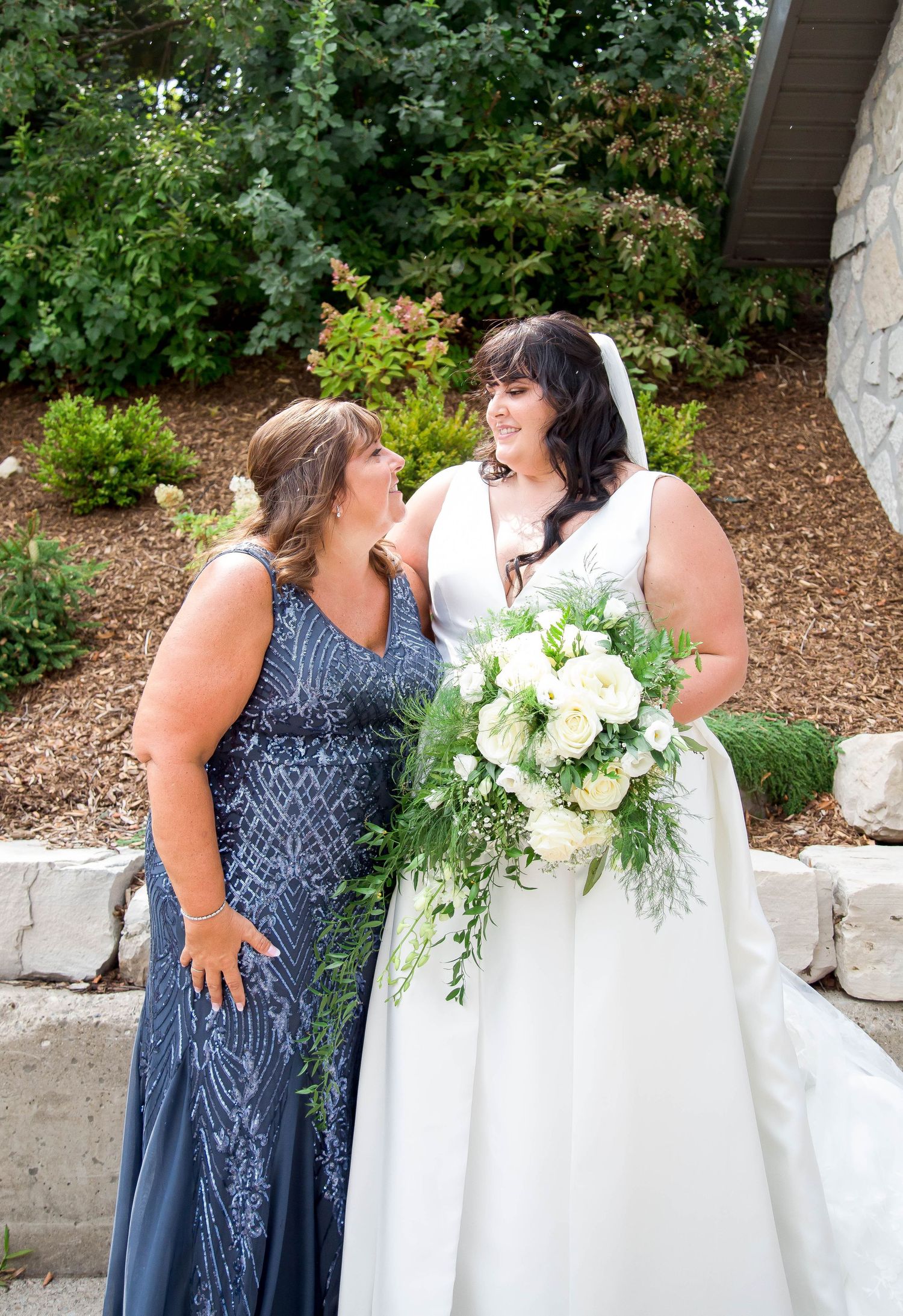 A bride in a white gown holding white roses smiles with her mother in a beaded navy gown.