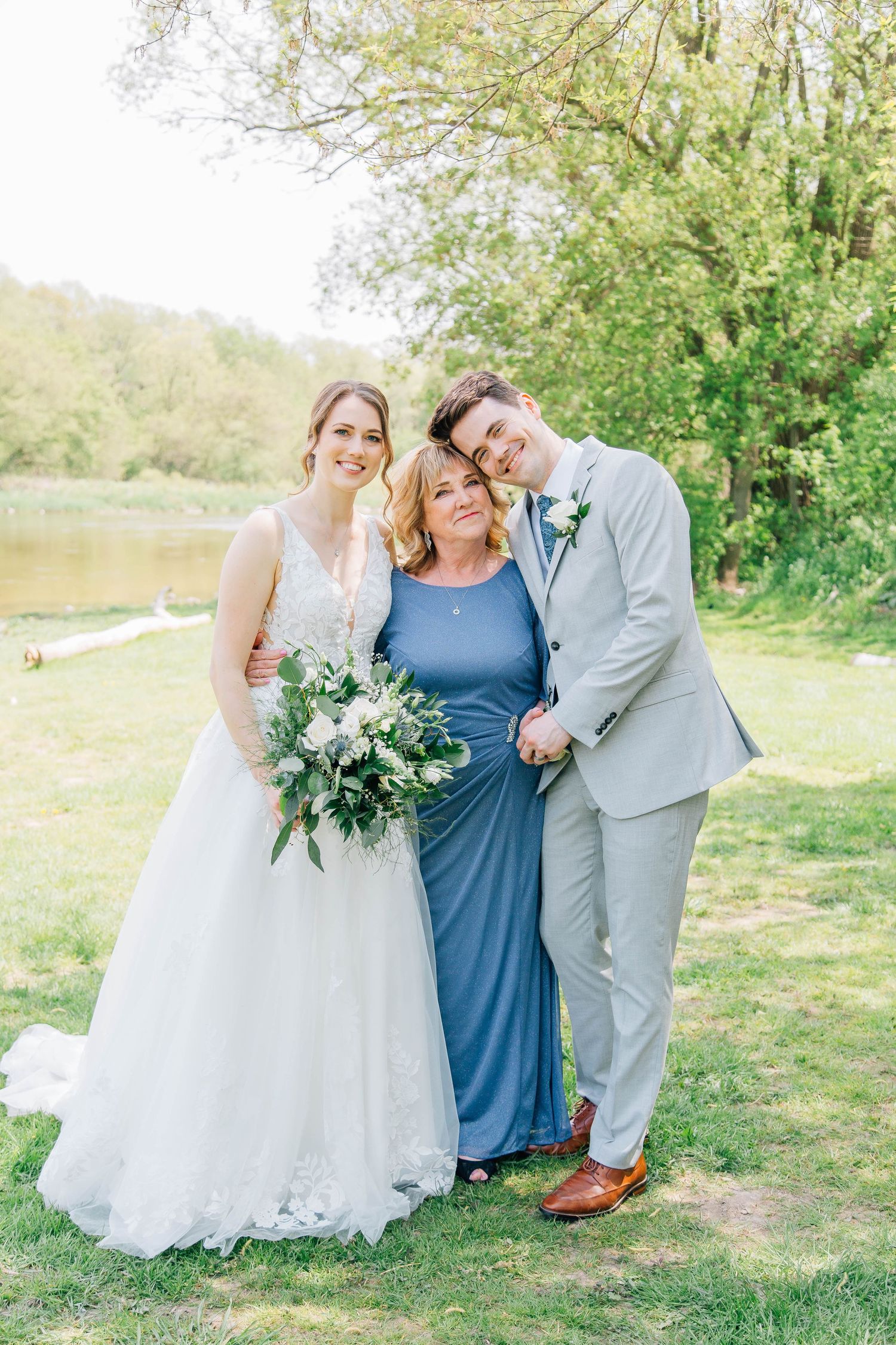 Bride and groom pose with mother in blue gown outdoors near a pond on their wedding day.