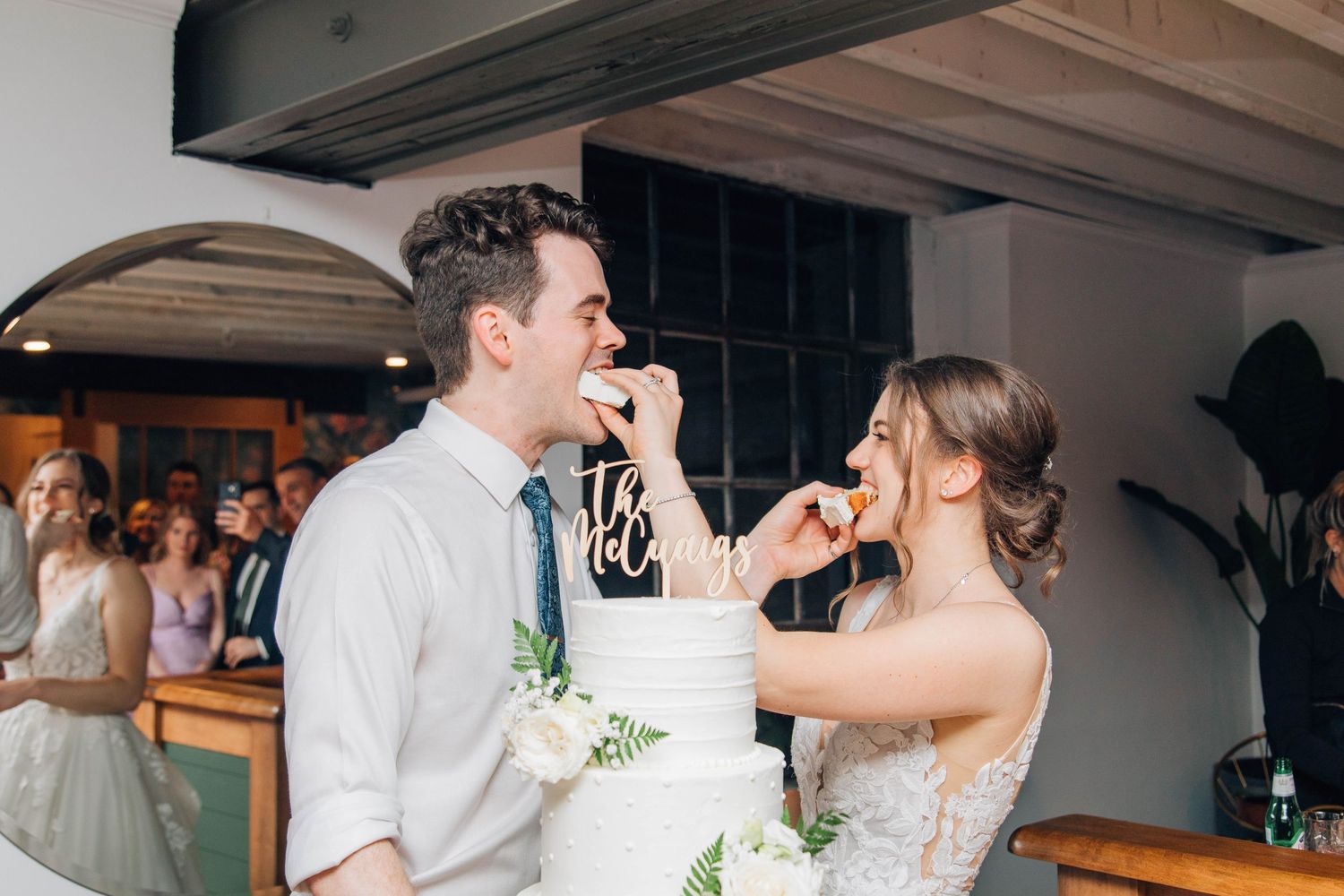 Bride and groom smiling while feeding each other wedding cake at their indoor reception celebration.