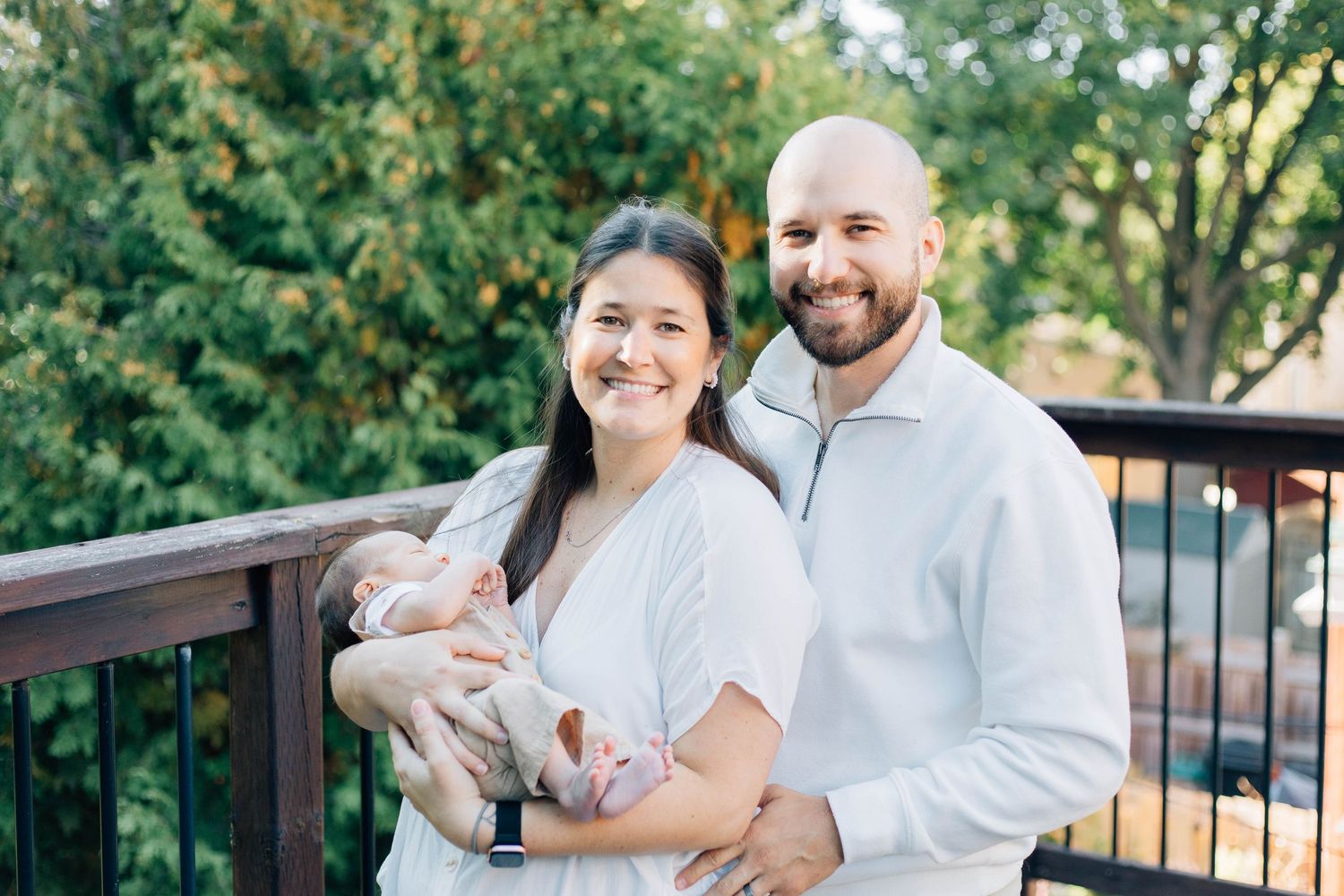 Smiling couple holding newborn baby on outdoor deck surrounded by green trees.