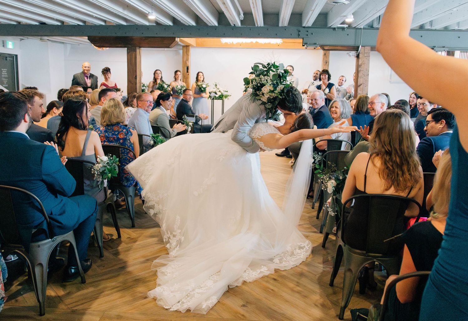 Newlyweds sharing romantic dip kiss while walking down aisle surrounded by cheering guests.