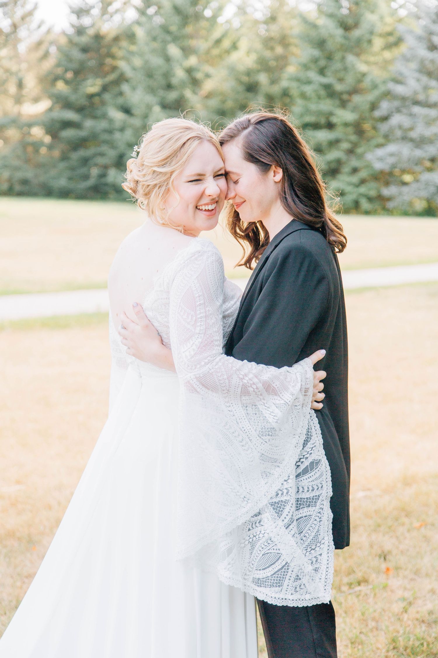 Two brides embrace and laugh together outdoors, one wearing a white lace gown and the other a black blazer at their wedding.