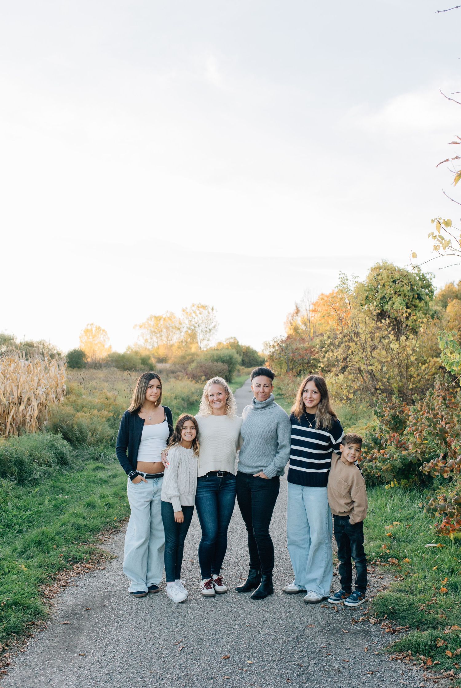 A family of six posing together on a gravel path surrounded by autumn foliage in natural light.