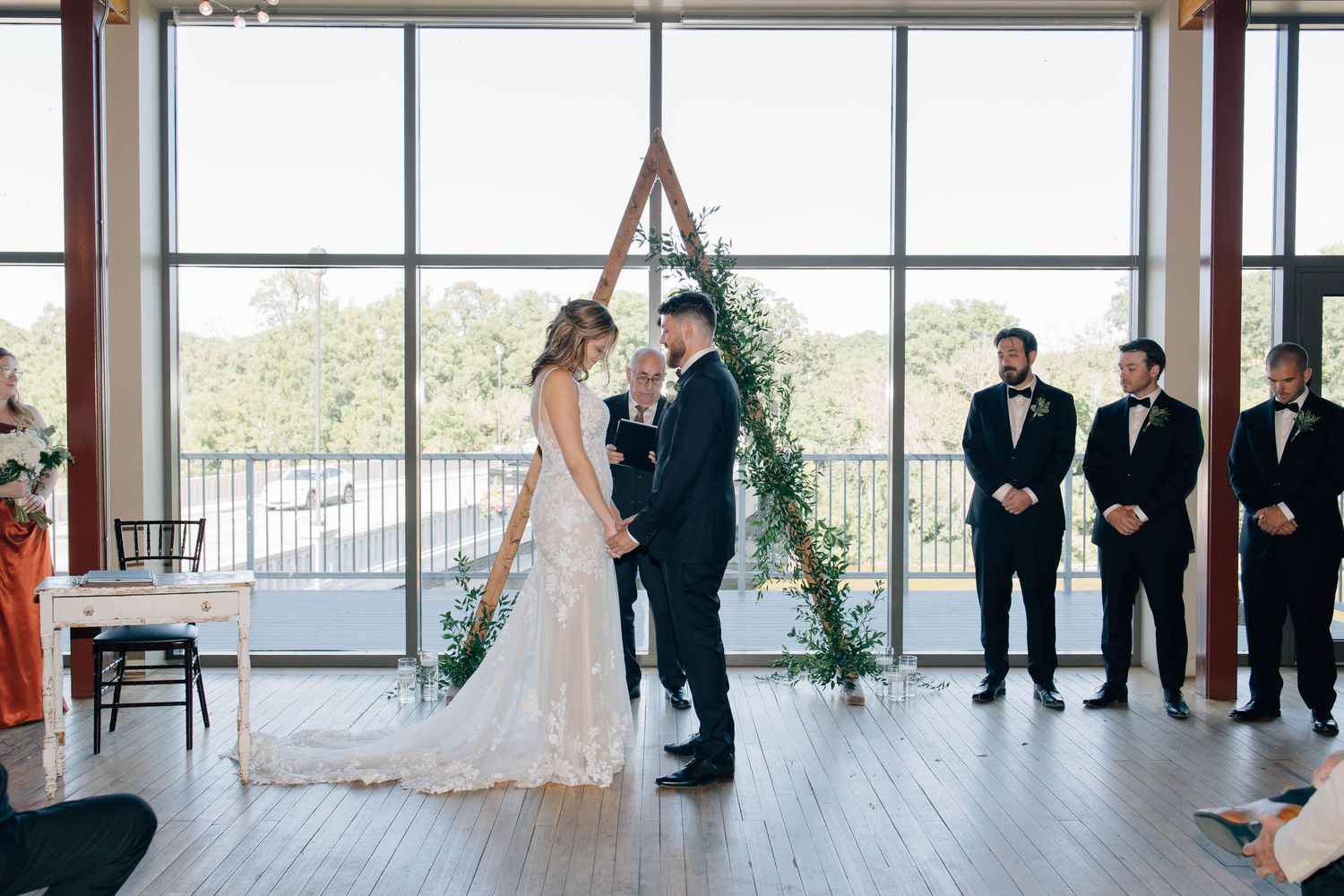 Bride and groom raising hands together beneath a triangular wooden arch during their indoor wedding ceremony.
