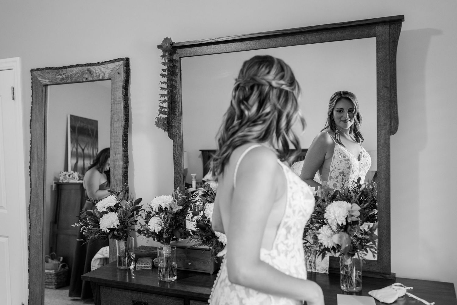 Black and white photo of bride in lace gown viewing her reflection in a large mirror.