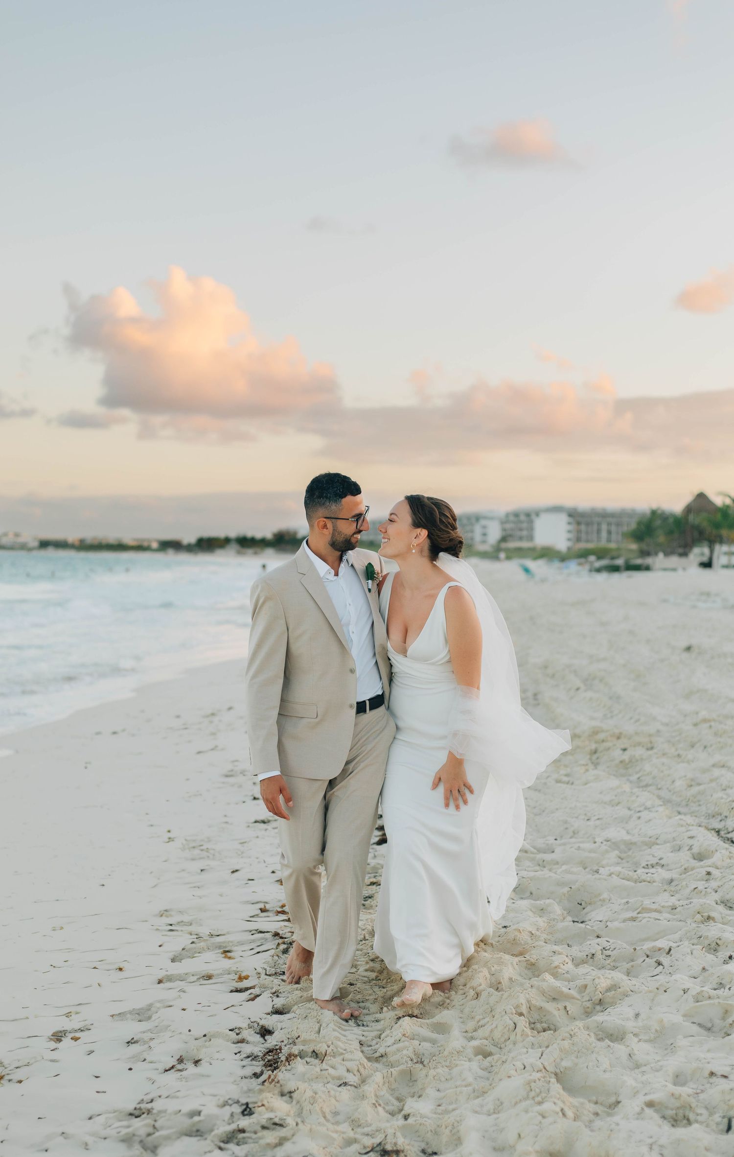 Bride and groom walking barefoot on a sandy beach at sunset with ocean waves in the background.