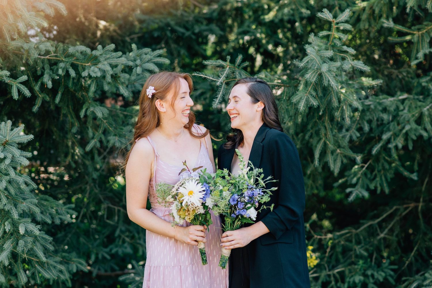 Two smiling women in pink and black dresses share a joyful moment holding colorful wildflower bouquets.