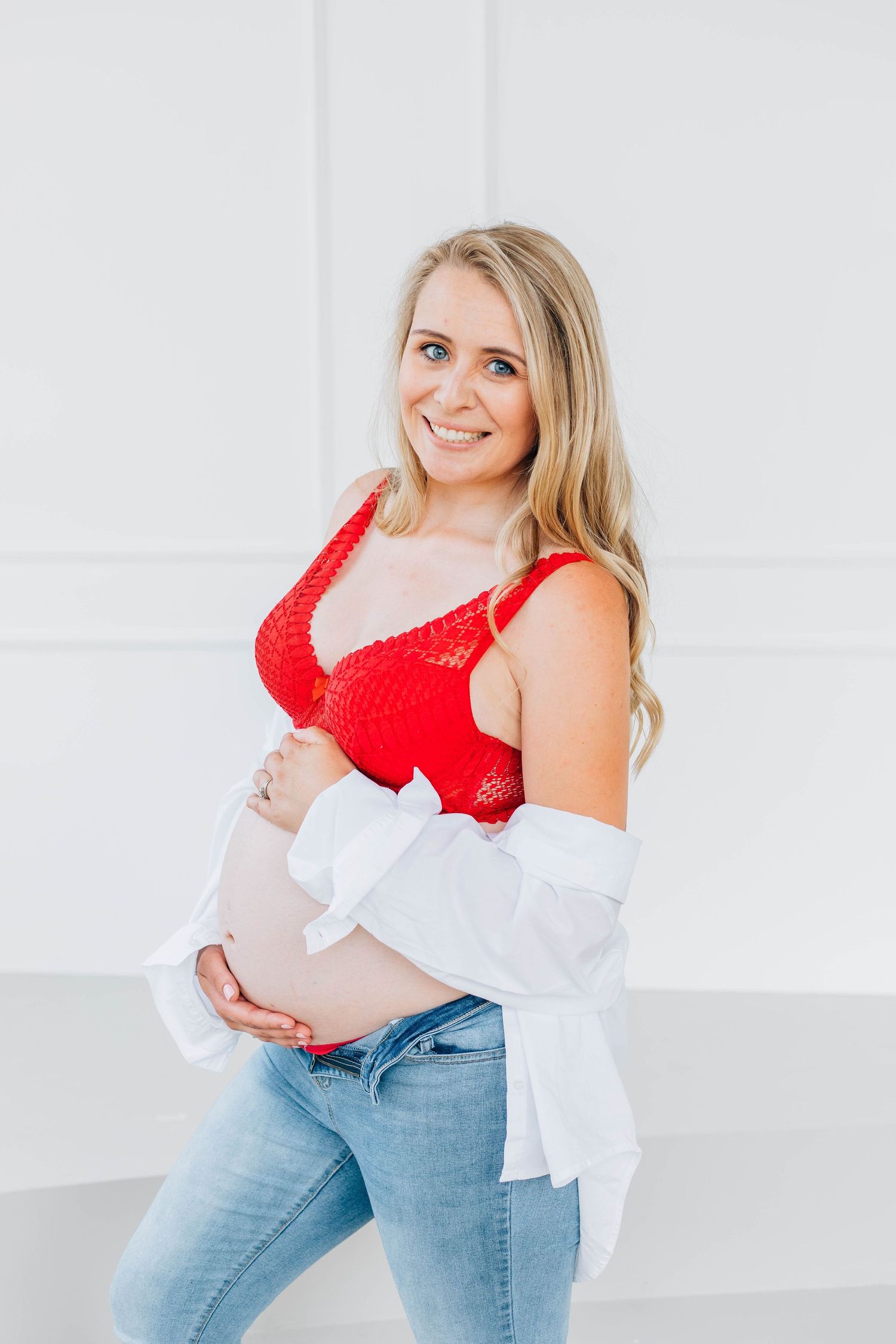 Smiling pregnant woman in red bralette and open white shirt cradling baby bump in white studio.