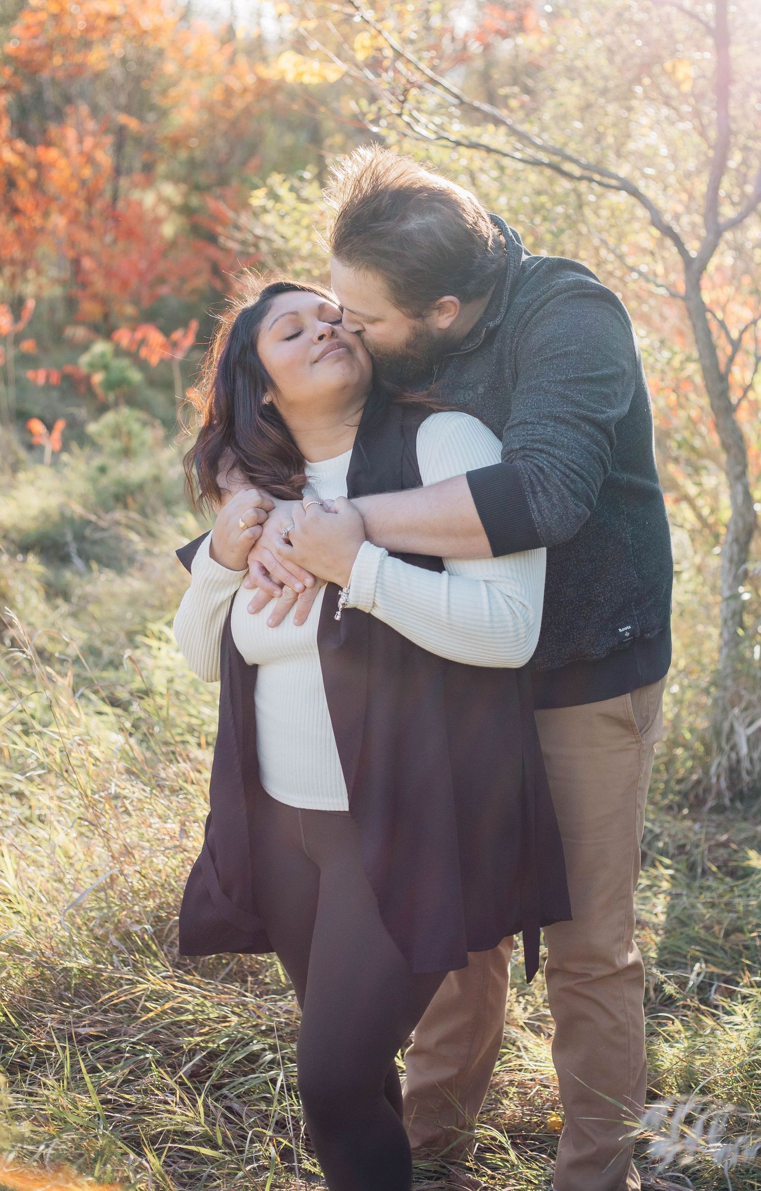 Bearded man tenderly kissing woman on cheek while embracing her in colorful autumn setting.