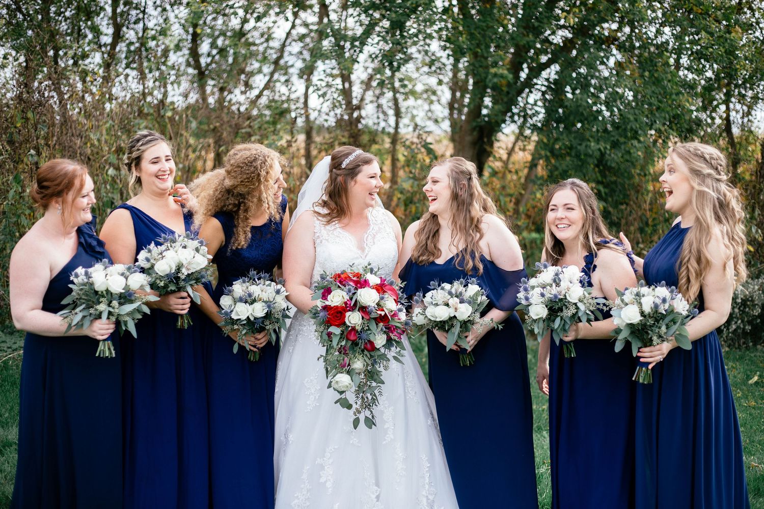 Bride and bridesmaids in navy dresses laugh together joyfully while holding floral bouquets outside.