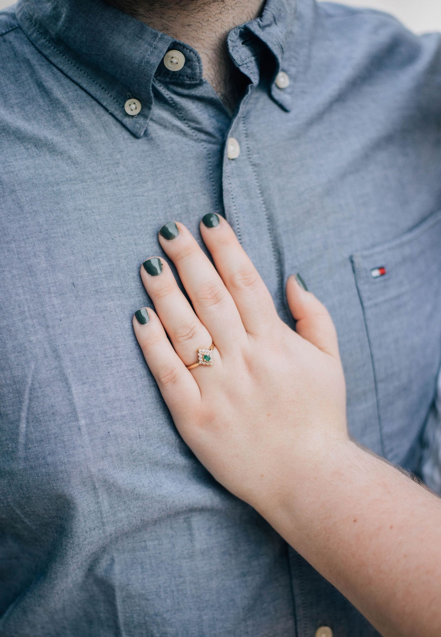 Close-up of womans hand with engagement ring and dark nail polish resting on mans blue Tommy Hilfiger shirt.