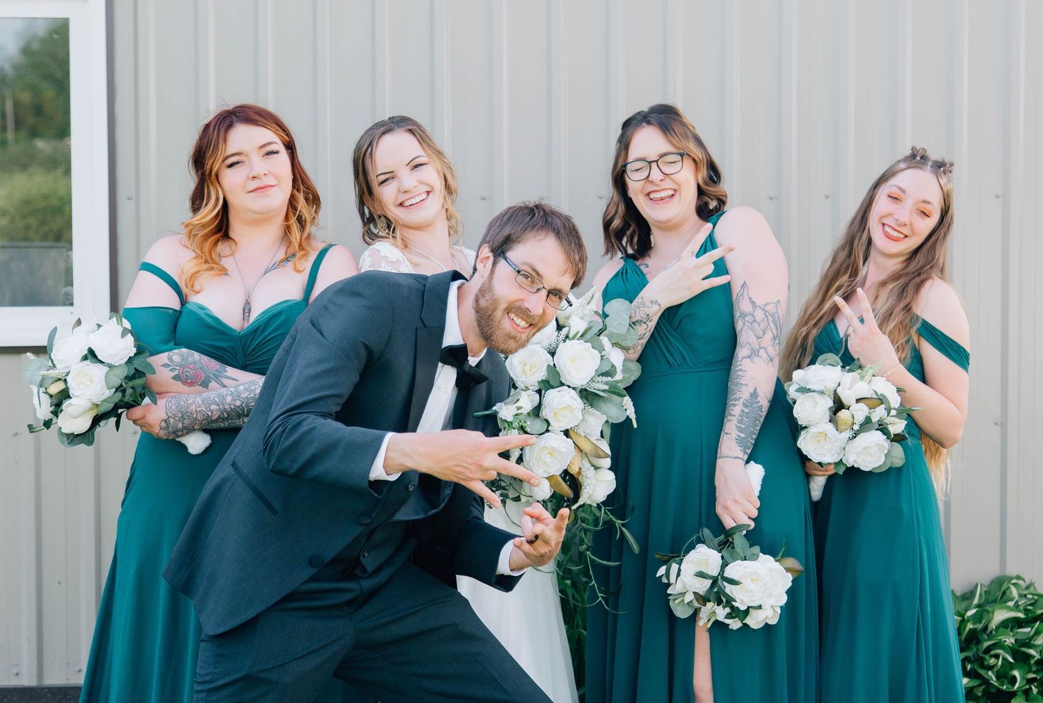 A groom poses playfully with four bridesmaids in emerald green dresses holding white floral bouquets outdoors.