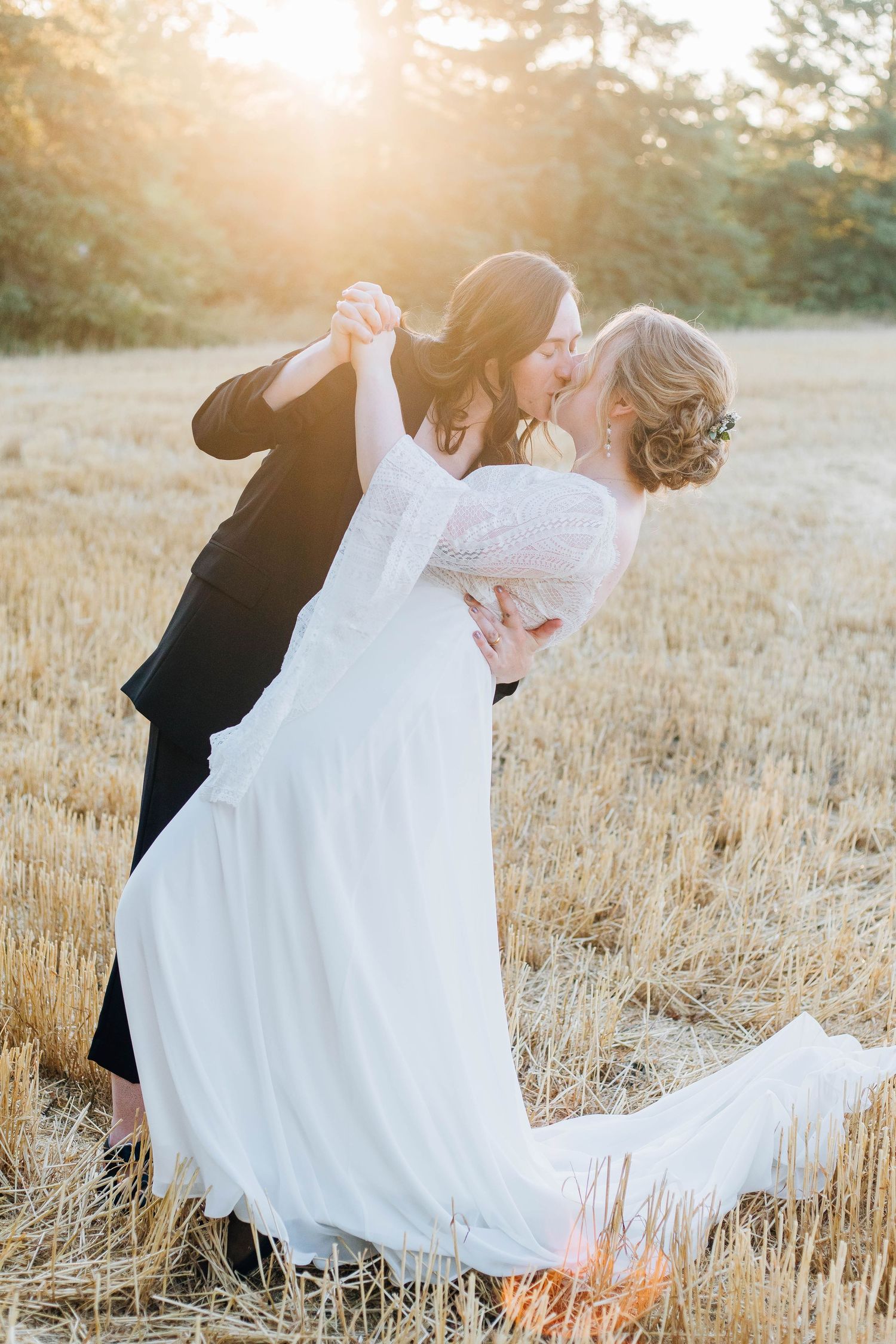Lesbian couple sharing a romantic dip kiss in a golden wheat field at sunset.