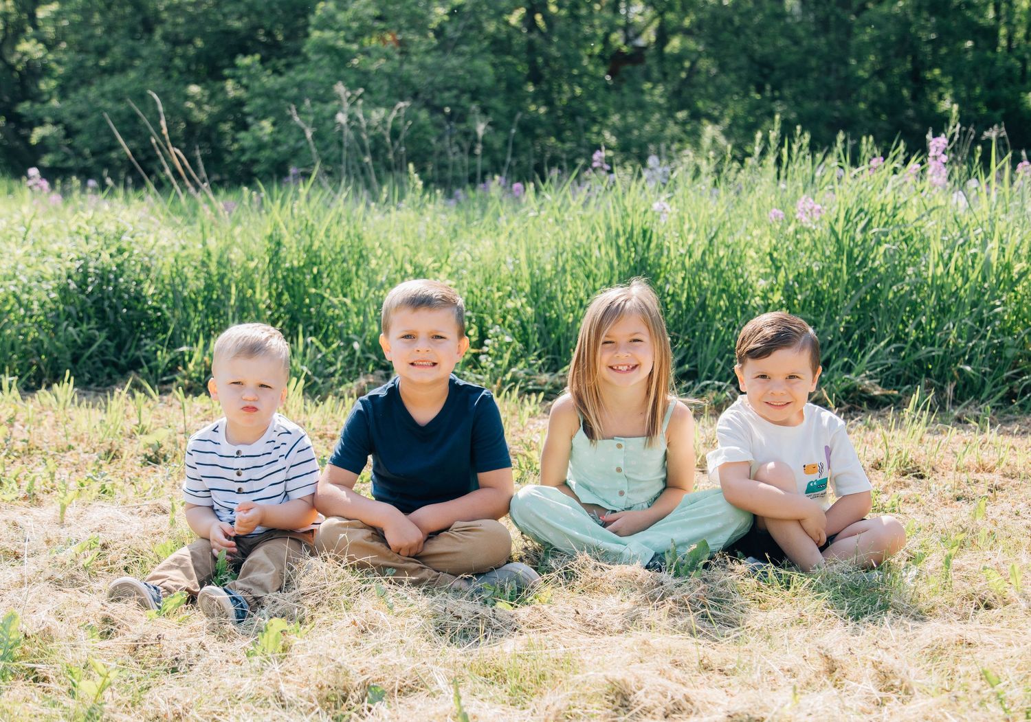 Four young children sitting together smiling in a sunny meadow with wildflowers behind them.