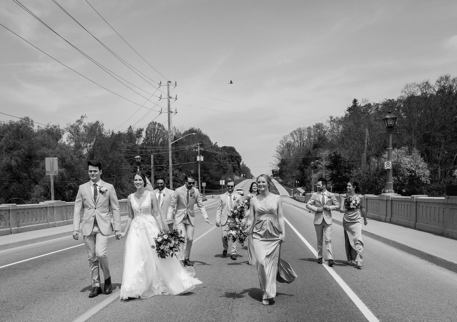 Black and white photo of wedding party walking across road bridge together on overcast spring day.