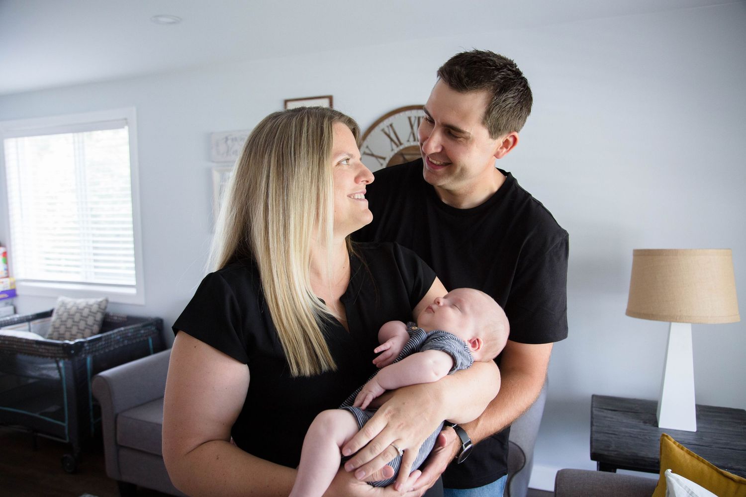 Happy couple in black shirts sharing a smile while holding newborn baby at home.