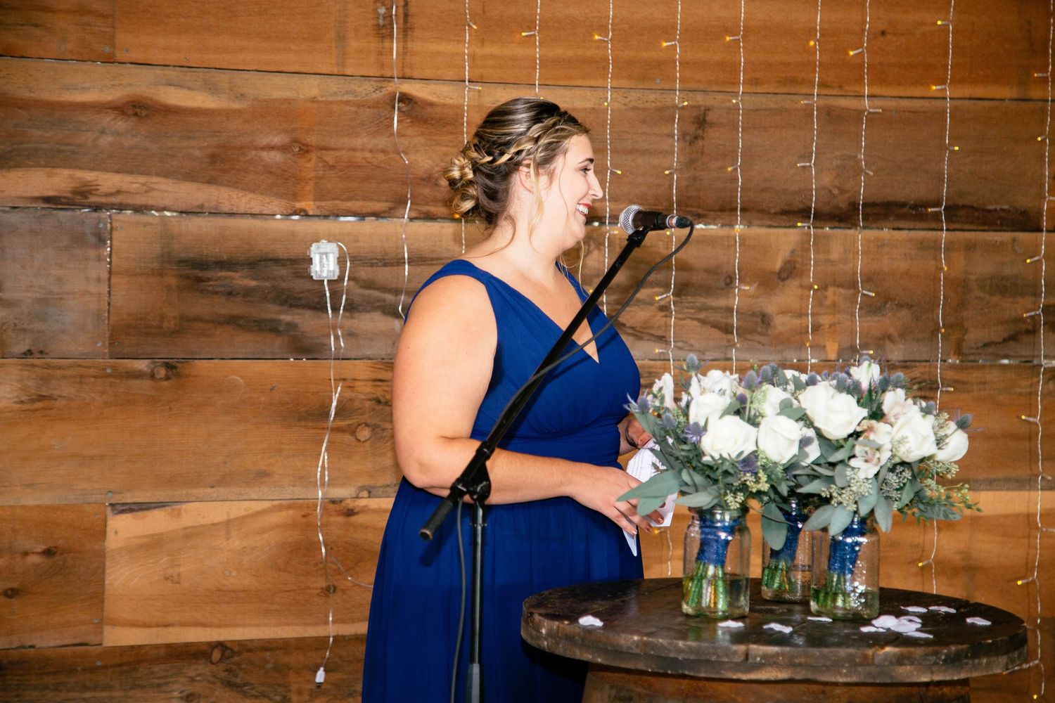 Woman in royal blue bridesmaid dress delivers speech at microphone during rustic barn wedding reception.