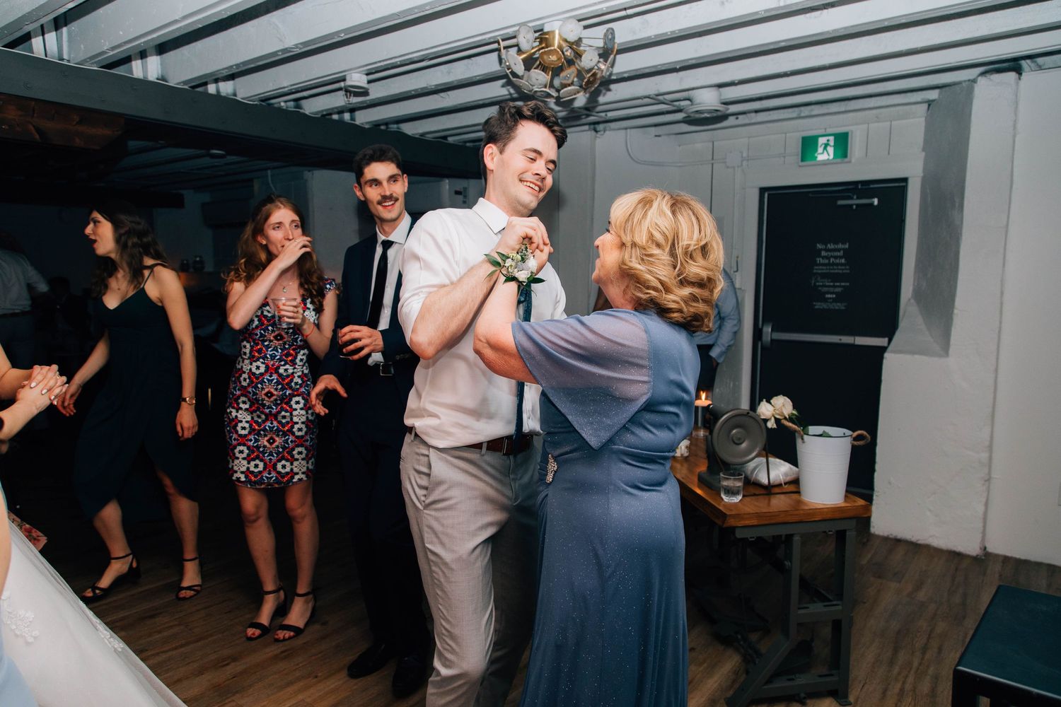 Groom in gray suit shares a dance with an older woman in blue at wedding reception.