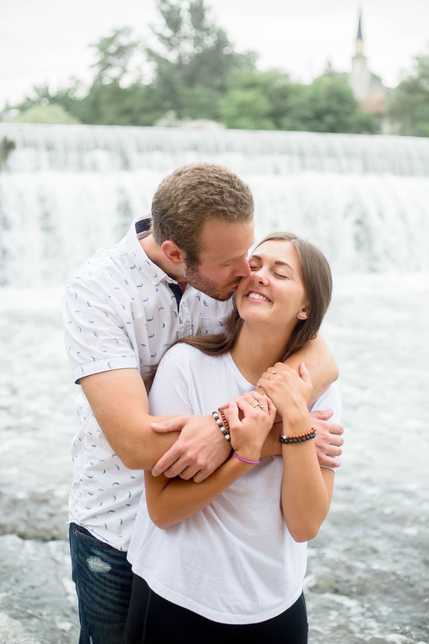 A man tenderly kisses a woman on the cheek while embracing her in front of a waterfall.