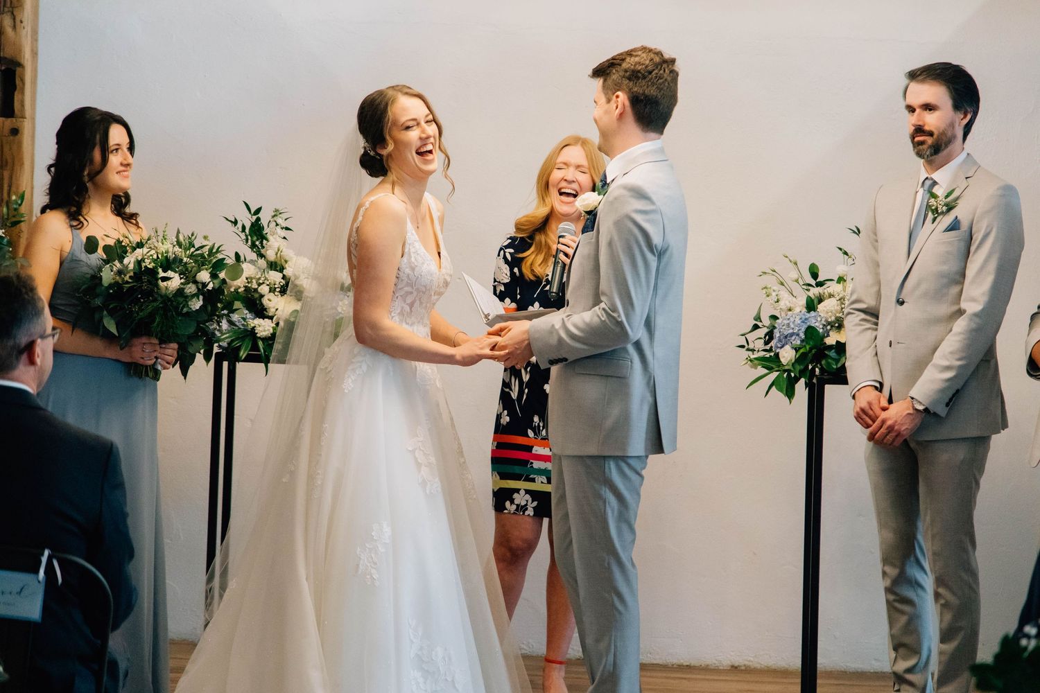 Bride and groom laughing joyfully during wedding ceremony with bridesmaids and officiant.