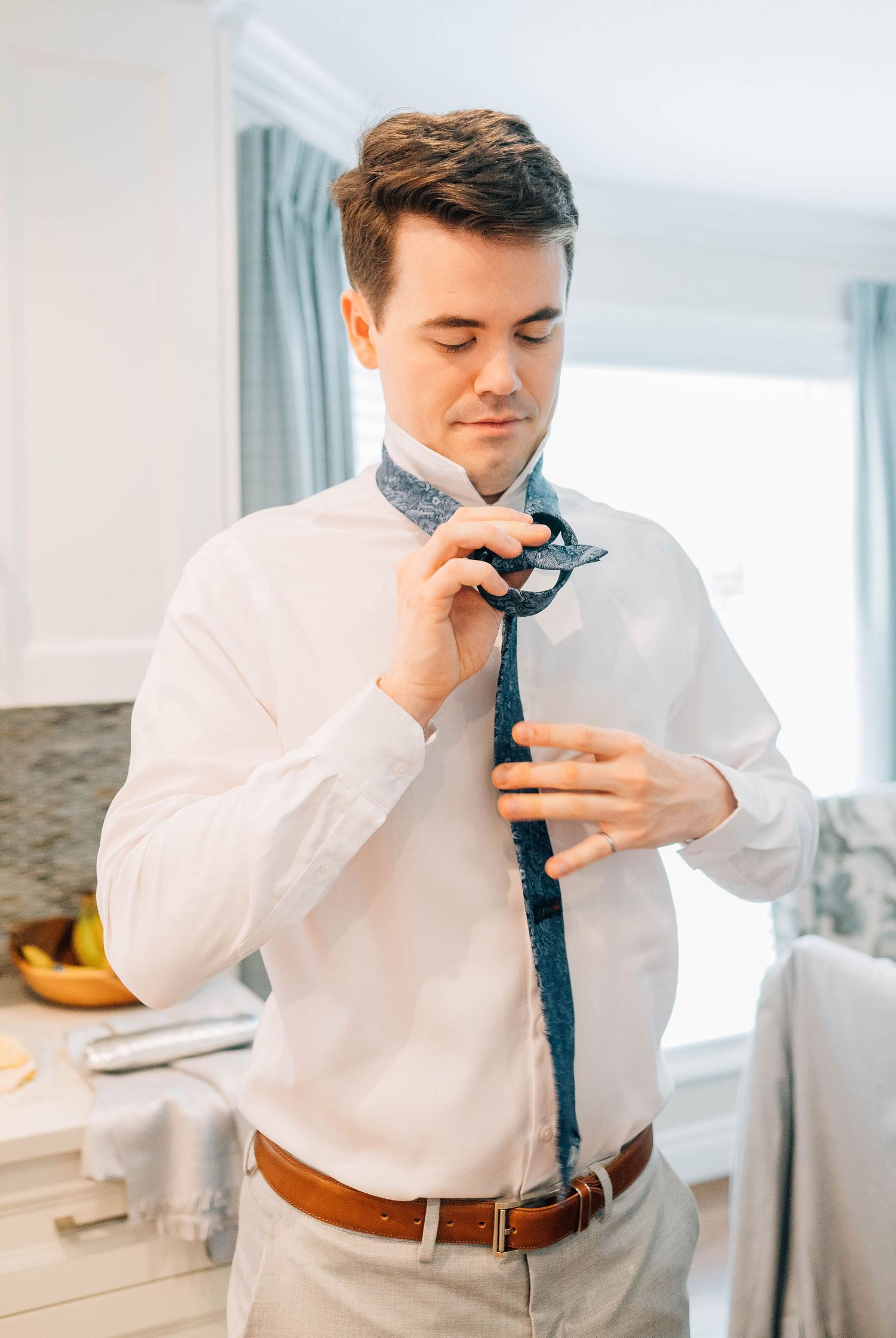 Groom in white dress shirt tying a blue floral tie while getting ready for his wedding day.