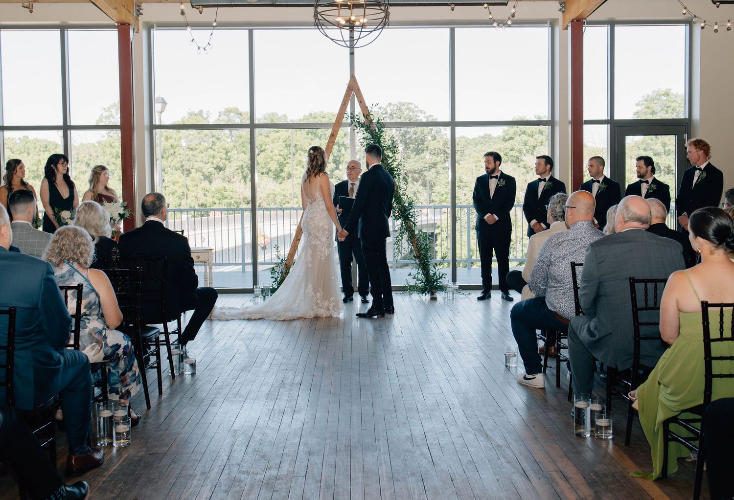 Wedding ceremony in a modern venue with large windows, guests seated as bride and groom exchange vows at a wooden arch.