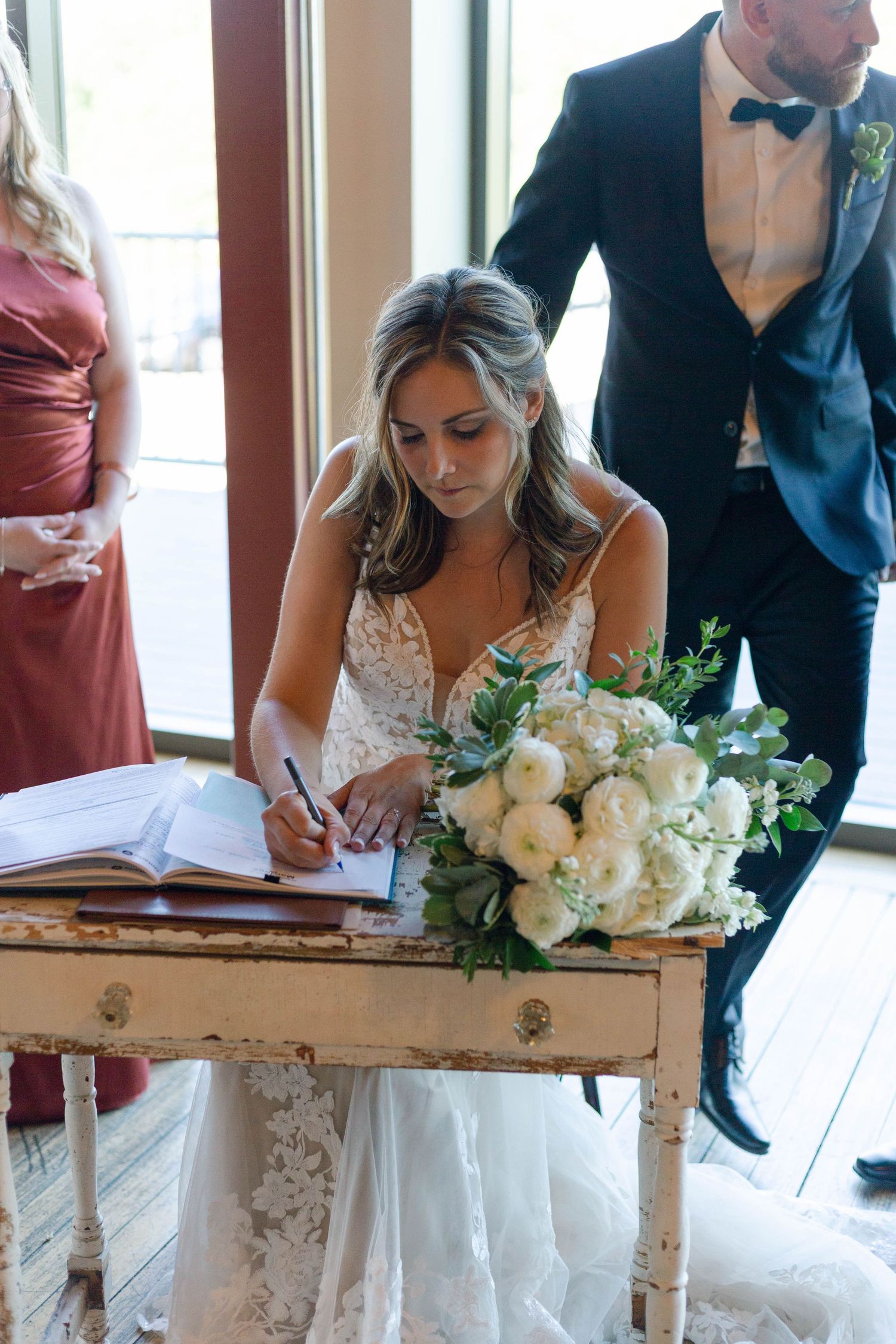 Bride in lace wedding gown signing marriage certificate at rustic white desk with white floral bouquet.