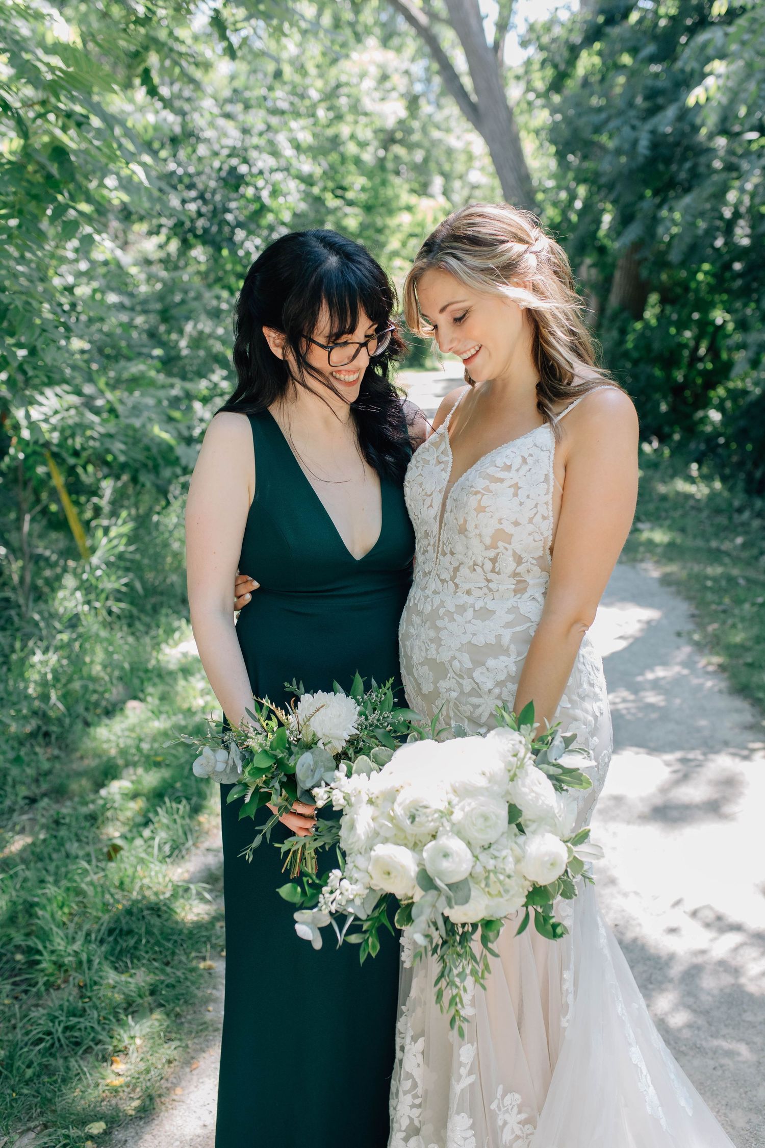 Bride in lace gown and bridesmaid in emerald dress share a tender moment holding white bouquets outdoors.