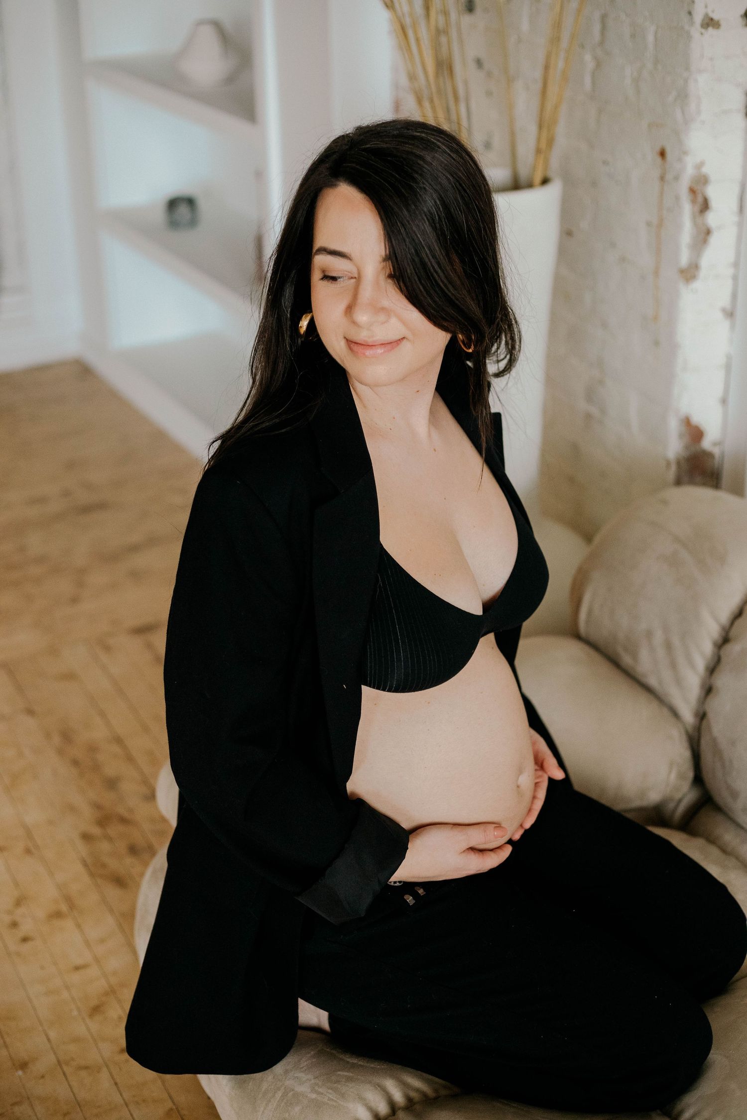 Smiling pregnant woman in black blazer and bra sitting on couch edge, lovingly cradling her baby bump.