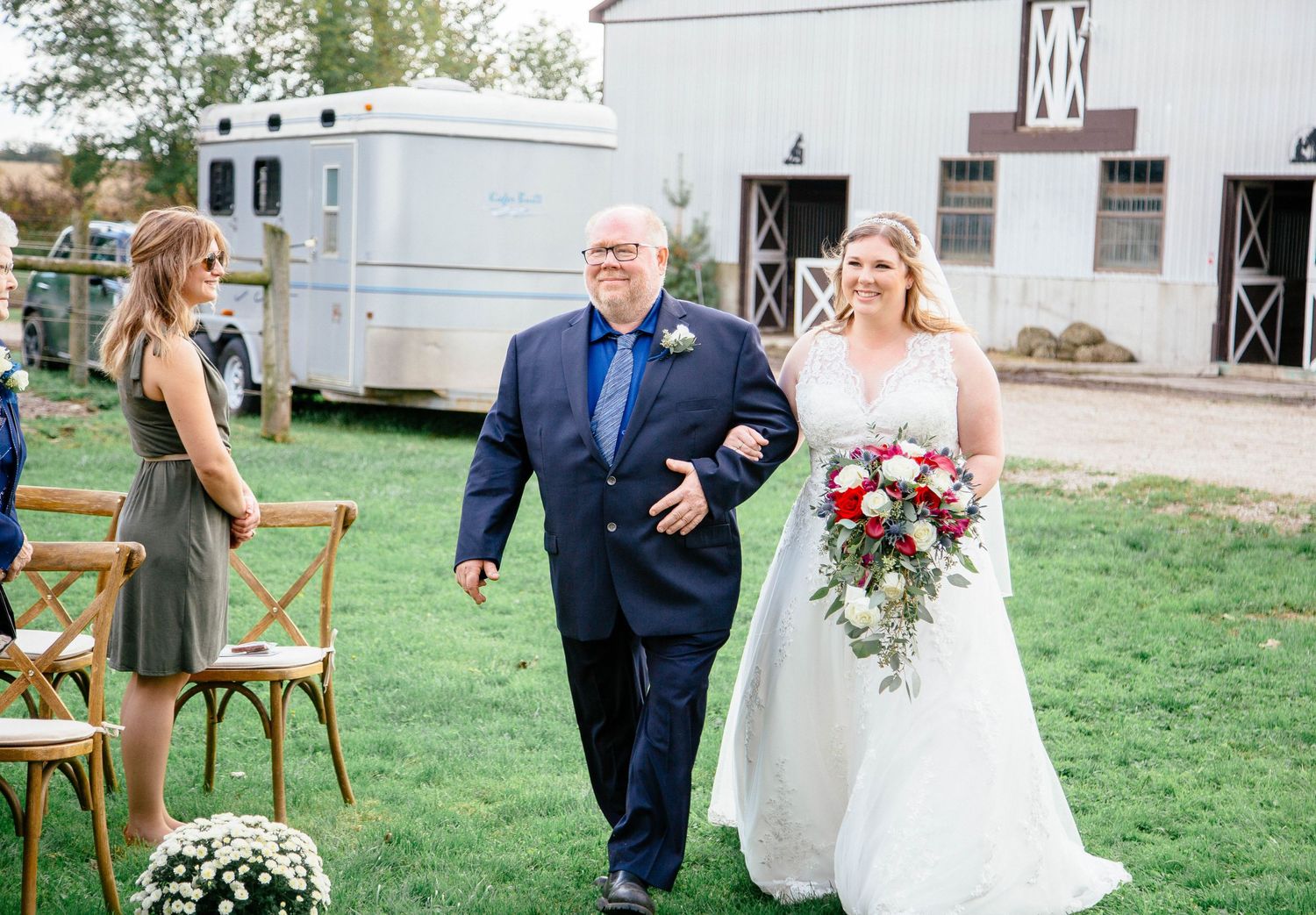 Bride in white gown walks down the aisle with her father at an outdoor barn wedding.