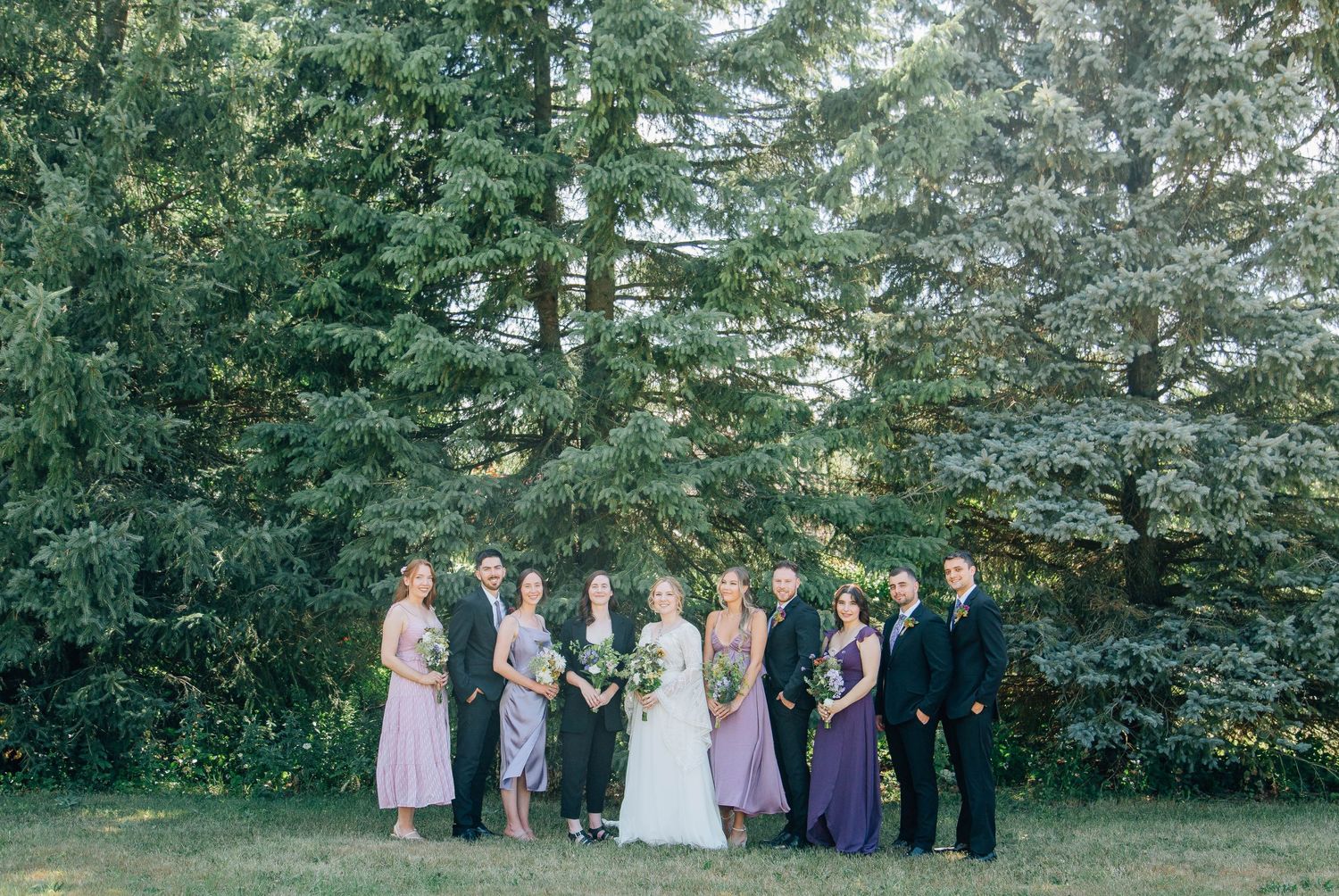 A wedding party of ten posing together in front of tall green trees outdoors.