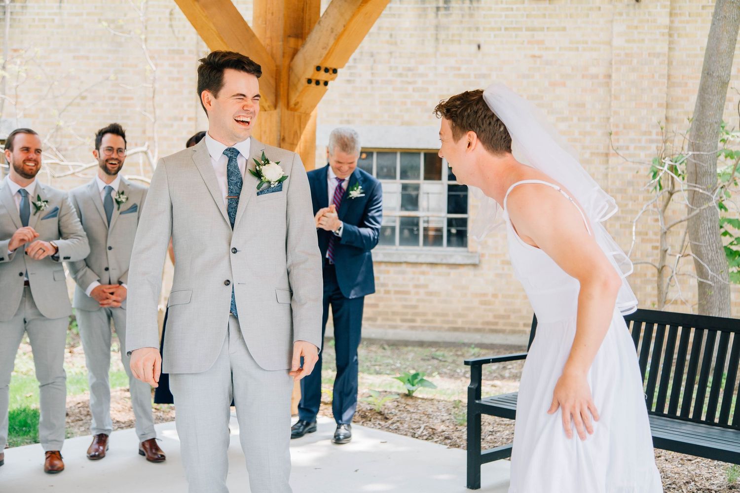 Groom laughing joyfully during first look moment with bride at outdoor wedding venue with groomsmen watching.